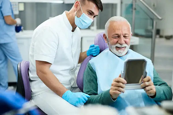 Happy, senior patient looking at himself in a mirror with his new smile at The Facial Center in Charleston, WV