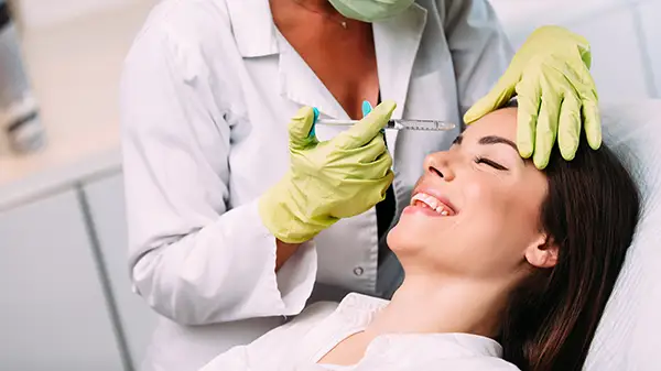 Smiling woman leaning back in a clinic chair, receiving Botox injection in her forehead to reduce wrinkles.